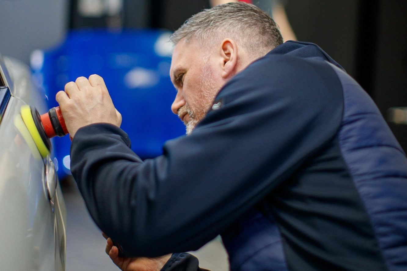 A man using a polishing tool on a car surface in a workshop setting.