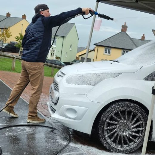A person using a pressure washer to clean a white car under a canopy.